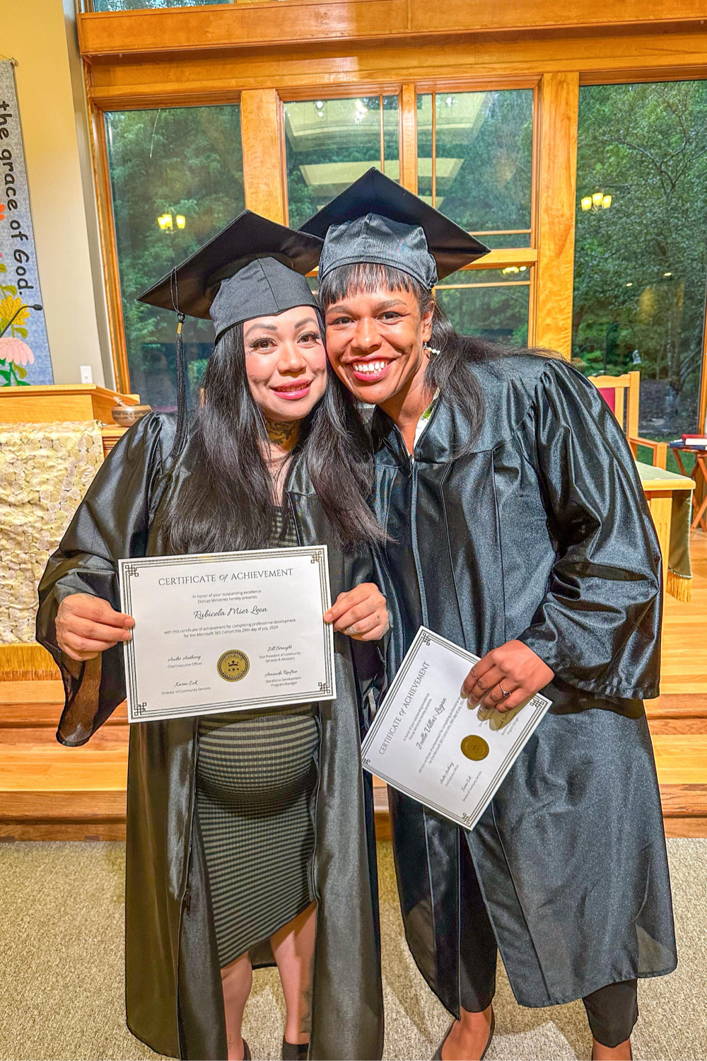 2 women smiling wearing a cap and gown and holding diplomas for a certificate of achievement.